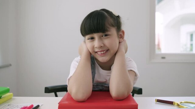 Happy cute Asian schoolgirl is sitting at a desk and looking at the webcam on a laptop computer while using a remote for online learning at home. Concept of new normal education in the homeschool. 