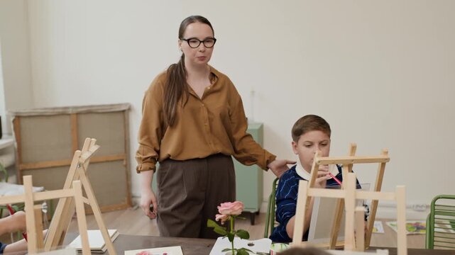 Caucasian female art teacher instructing children during drawing master class at easels. Footage suitable for promoting creative child education and artistic workshop campaigns
