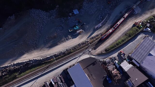 Aerial view of the red Bernina Express train traveling along tracks past industrial buildings and construction sites in Brusio, Grisons, Switzerland.