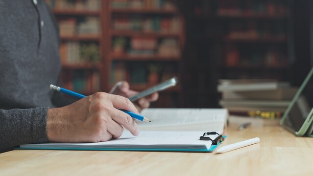 Focused student engages in serious study at library desk, balancing study routines with phone research as academic study advances knowledge.