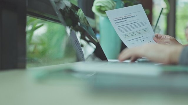 Hands conduct audit review decarbonization audit paperwork beside laptop computer, completing corporate audit analysis alongside sustainability documentation.