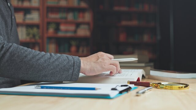 Focused researcher conducts literature research at desk, cross referencing research material beside academic textbook in library.