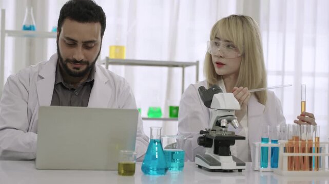 Team of happy professional scientists and researchers in lab coats using microscope and a laptop while conducting chemistry experiments with liquid in test tubes and beakers in the science laborator