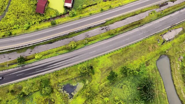 Aerial view of Tanjung Mulia highway surrounded by lush green vegetation, red-roofed buildings, and a vibrant green pond in Medan Deli, North Sumatra, Indonesia.