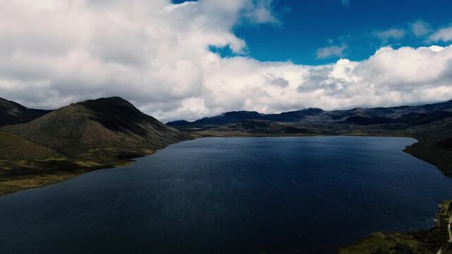 Aerial view over tussock grasses and low shrubs covering rolling slopes, with layered mountain ridges beneath a blue sky and large white clouds. Drone footage of the Andean highlands in Ecuador captur