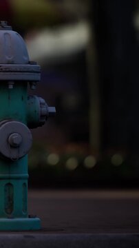A fire hydrant is located on the side of a street in a busy city. The light is fading, and the area shows some colors in the background. This moment captures an everyday scene.