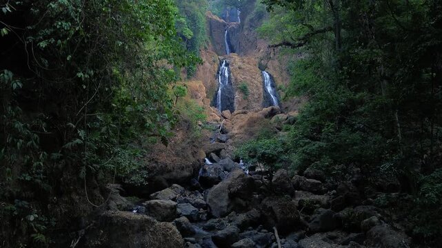 Aerial drone footage of a scenic waterfall flowing over rocky terrain through a dense tropical forest. Multiple cascading streams descend between mossy rocks and lush green vegetation.