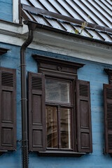 A timber window with louvered shutters sits below a metal roof and a plant filled gutter, showing facade joinery and rainwater drainage components for building maintenance © K