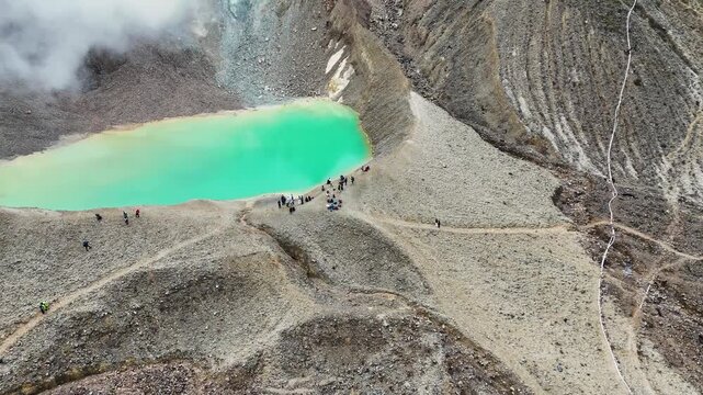 Aerial view of Mount Papandayan featuring a vibrant turquoise volcanic lake with people gathered on the rocky shore in Garut, West Java, Indonesia.