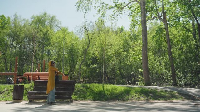 Woman wearing yellow coat stands near wooden benches with black trash bins in urban green space. Woman interacts with her bag placing it on and picking up from bench as woman moves