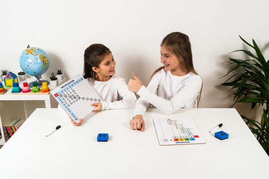 Two schoolgirls studying mathematics together at a table, with one girl holding a multiplication table board while her friend gives a thumbs-up gesture of encouragement.