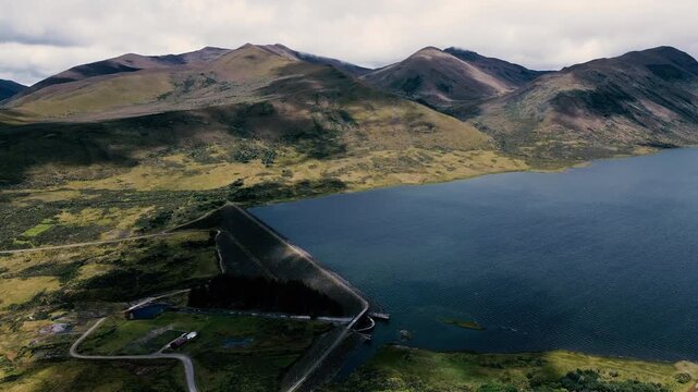 A pale unpaved road curves across open, sparsely vegetated highland terrain, with dark mountain ridges on the horizon and scattered patches of trees and water below a cloud-filled sky. Aerial drone fo