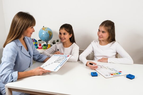 In a bright classroom, a teacher assists two girls with math concepts using educational boards. The kids are focused and involved, making learning enjoyable and interactive with visual tools.