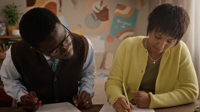 African American tutor helping Black teenage girl solve exam tasks on paper sheets at desk. Video suitable for private tutoring, test prep courses, and education campaigns