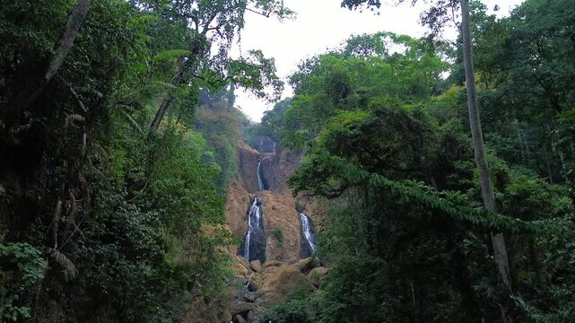 Aerial drone footage of a scenic waterfall flowing over rocky terrain through a dense tropical forest. Multiple cascading streams descend between mossy rocks and lush green vegetation.