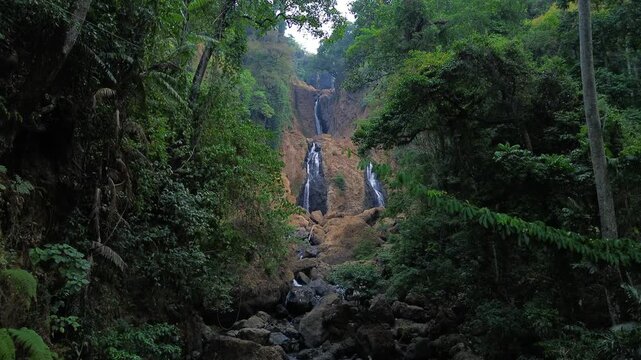 Aerial drone footage of a scenic waterfall flowing over rocky terrain through a dense tropical forest. Multiple cascading streams descend between mossy rocks and lush green vegetation.