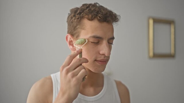 Young man rolling jade roller over cheek and nose with closed eyes in studio; self care relaxation.