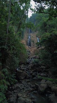 Aerial drone footage of a scenic waterfall flowing over rocky terrain through a dense tropical forest. Multiple cascading streams descend between mossy rocks and lush green vegetation.