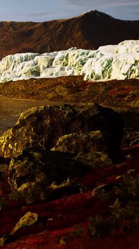 A glacier is seen in the background with large ice formations reflecting light. In the foreground, there are dark rocks and some red vegetation. This scene takes place during sunset in a remote area.