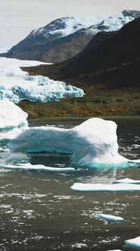 Large icebergs in calm water with mountains in the background. The sun is shining and the sky is clear. The water reflects the ice and mountains.