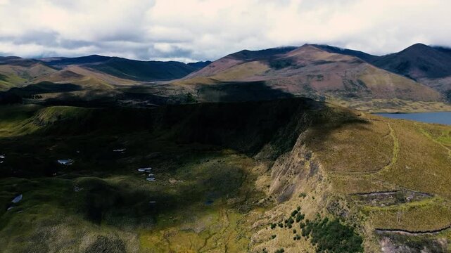 Aerial drone view over an Andean valley in Ecuador, where a pale winding road crosses rolling highland hills beside a narrow stream. Sparse grass and low vegetation cover the rugged slopes, with layer