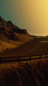 A winding road stretches through an arid landscape with rocky mountains in the background. The sky shows shades of orange and blue during sunset.