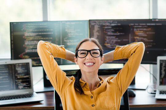 Smiling female software developer relaxed at her desk with multiple computer screens showing code in a modern tech office environment
