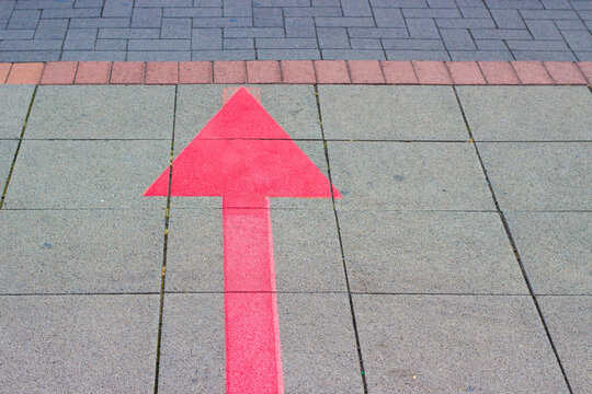 Bright pink directional arrow painted on large gray pavement tiles with red and dark brick bands visible in the upper background, outdoor ground marking