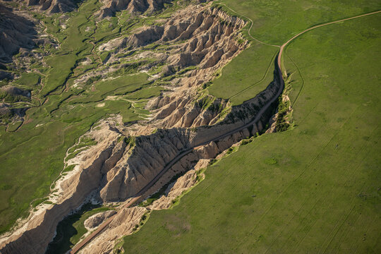 Aerial view of Badlands National Park showing eroded rock formations and deep canyons contrasting with green prairie grasslands and a winding road in Interior, South Dakota, United States.