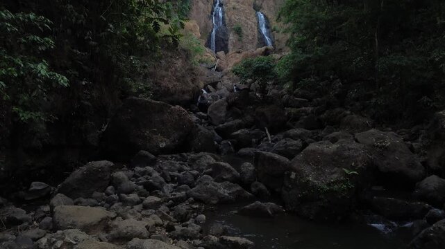 Aerial drone footage of a scenic waterfall flowing over rocky terrain through a dense tropical forest. Multiple cascading streams descend between mossy rocks and lush green vegetation.