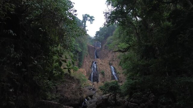 Aerial drone footage of a scenic waterfall flowing over rocky terrain through a dense tropical forest. Multiple cascading streams descend between mossy rocks and lush green vegetation.