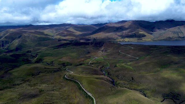 Aerial view of open Andean highland grassland in Ecuador, with a narrow winding stream crossing a broad plateau beneath rugged mountain ridges. Large clouds cast shadows across the green-brown terrain
