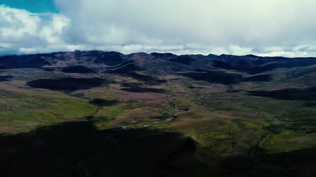 A winding road cuts across open highland terrain beneath layered mountain ridges, with a narrow stream and a lake visible in the valleys. Drone footage in the Andes of Ecuador captures green-brown hil