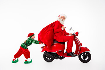 Fototapeta premium Santa Claus on a red scooter carrying a large gift sack while a playful elf child pushes in a whimsical Christmas holiday scene