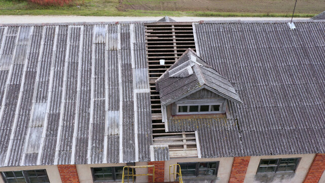 Aerial view of a damaged roof with missing corrugated panels revealing the wooden frame and a dormer window on a brick building in Latvija.