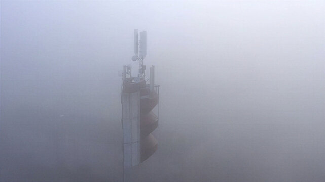 Aerial view of a telecommunications tower emerging from dense fog with antennas and equipment visible at the top in Latvija.