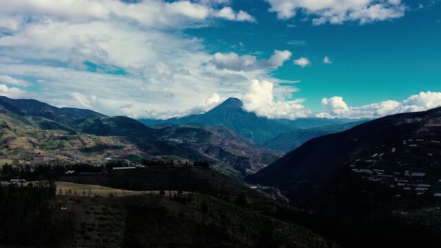 Aerial drone view of a narrow meandering stream crossing open green and brown grassland below rugged mountain ridges. Cloud cover and broad shadows shape the expansive landscape in the Ecuadorian Ande