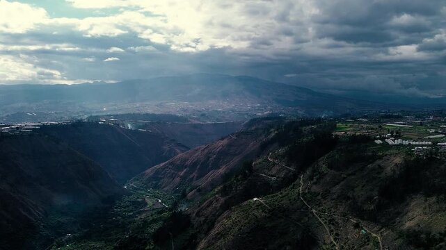 Drone footage looks across a broad Andean valley in Ecuador, where cultivated hillsides and scattered rural structures frame layers of rugged mountains. A prominent peak rises beneath billowing white 