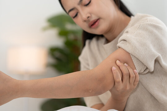 Young Asian woman suffering from elbow pain, holding arm with discomfort while sitting on bed at home, muscle strain, injury, healthcare problem and body pain concept in bedroom