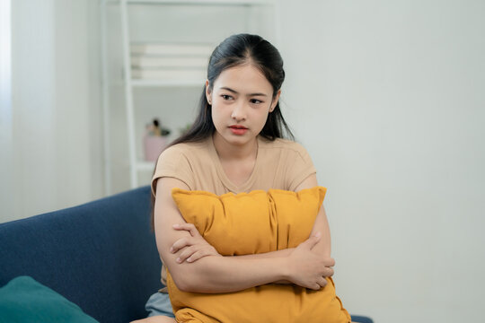 Asian woman feeling anxious and lonely hugging pillow on sofa at home, mental health, depression, emotional stress, sadness and overthinking concept, lifestyle problem indoors