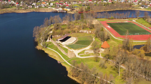 Aerial view of the historic Aluksne Castle ruins, featuring stone walls, a conical roof tower, an outdoor stage, and a soccer field, on the shore of a lake in Latvija.