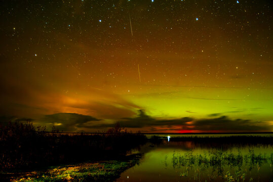 A spectacular night sky with the aurora borealis over a calm lake, clouds shimmering in greenish and golden light, and a dark shoreline with a small wooden pier.
