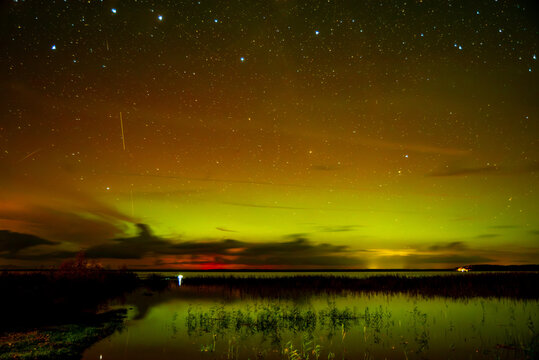 A spectacular night sky with the aurora borealis over a calm lake, clouds shimmering in greenish and golden light, and a dark shoreline with a small wooden pier.
