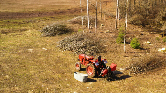 Aerial view of a red tractor parked in a field with piles of cut branches and trees in Latvija.