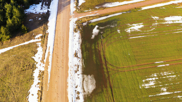 Aerial view of a dirt road cutting through rural fields with patches of snow and tire tracks in Latvia.