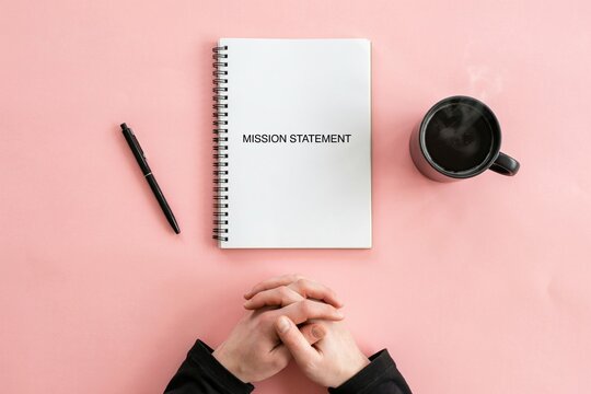 A person sitting at a pink desk with folded hands next to a notebook with the text mission statement.