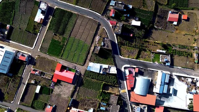 Drone footage looks across cultivated fields, roads and a settlement in a broad Andean valley, with steep green mountains and a prominent volcanic peak beneath scattered clouds. The landscape is in Tu