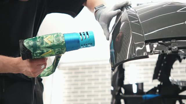 Detailer using a heat gun tool on a car bumper for final stage PPF installation, heating to securely fix edges activate the adhesive layer. Male technician completes paint protection film wrapping. 