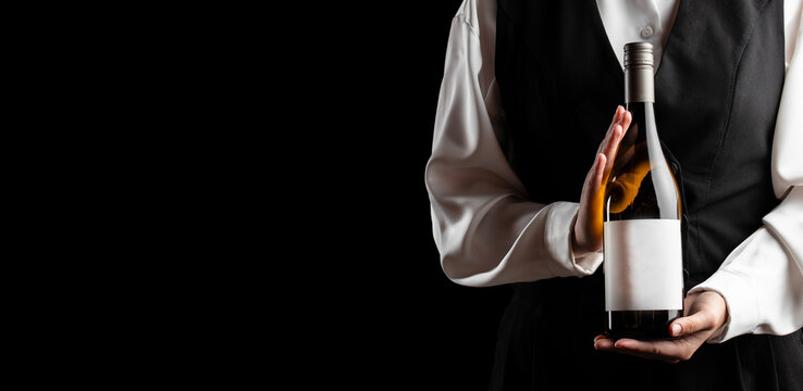Server holding an unbranded wine bottle with blank label against a black background, cropped formal uniform and isolated restaurant service presentation