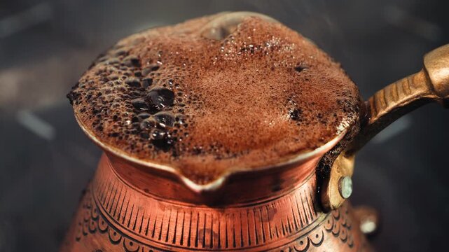 Close-up of Turkish coffee foam boiling and overflowing inside a traditional ornate copper cezve, with a hand quickly removing it from the stove. . High quality 4k footage